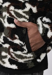 Fleecejakke med camouflage-mønster i grøn, brun, sort og hvid. Har en sidelomme og tekstureret, fluffy overflade.