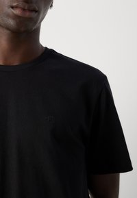 Close-up van een persoon die een eenvoudige zwarte t-shirt met korte mouwen draagt, met een klein, subtiel "cK" logo op de linkerborst.