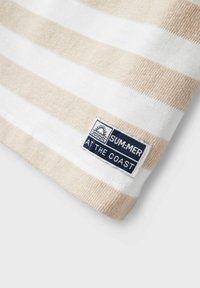 Tela de algodón a rayas en beige y blanco, con una superficie texturizada. En el borde se puede ver una etiqueta rectangular con texto azul y un ícono.