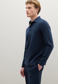 Marineblaues langärmeliges Poloshirt mit Kragen und Knopfleiste, aus glattem Stoff. Kombiniert mit passenden Hosen mit seitlicher Reißverschlusstasche.
