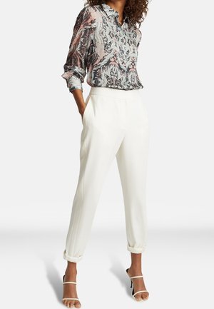 Kobieta ubrana w długą koszulę z wzorem, wsuniętą w białe, wysokowaste, zwężane spodnie z podwiniętymi nogawkami oraz beżowe sandały na obcasie.