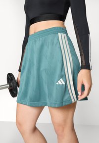Teal színű sport szoknya texturált mintával, fehér háromsávos díszítéssel és hímzett logóval az alsó szegélyen.