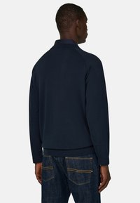 Homme portant une chemise à manches longues bleu marine foncé et un jean bleu, debout de dos sur un fond clair uni.