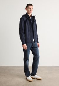 Marineblauwe ritssluitingjack met capuchon, voorzien van een gladde textuur. Draag het met donkerblauwe jeans en witte schoenen met gumzolen. Eenvoudig, casual ontwerp.