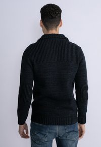 Pulover tricotat închis la culoare, cu guler ribbat și detalii pe umeri, având pe elbow-uri petice texturate și manșete ajustate. Purtați cu blugi blue deschis.
