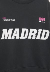 Čierna mikina s veľkým bielym textom "MADRID," ružovým "1991," a menším "nový KREATÍVNY TÍM." Mäkká textúra, okrúhly výstrih, neformálny dizajn.