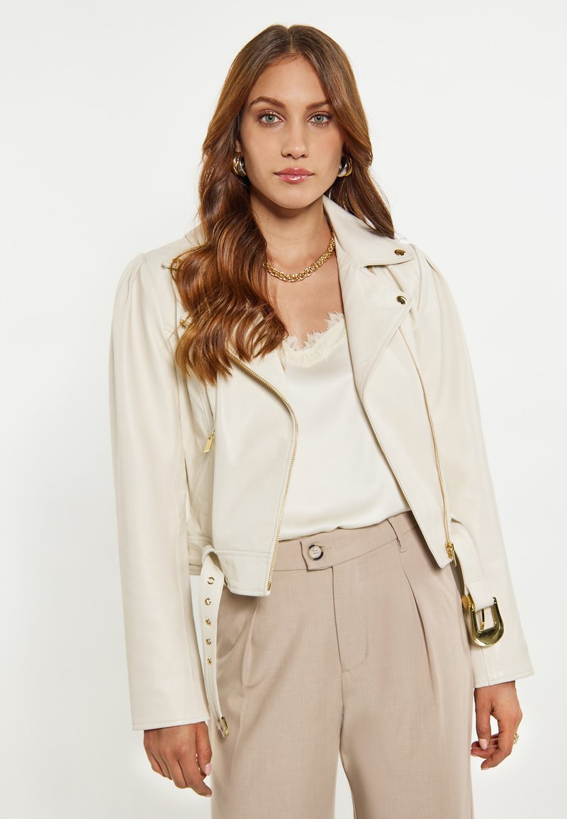 Giacca da motociclista in pelle crema con hardware metallico, chiusure con zip e fibbie sui lati. Indossata su un top di raso chiaro e pantaloni beige plissettati.