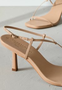 Beige sandalen met hakken en banden, met een vierkante neus en blokhak. Gemaakt van glad materiaal met een minimalistisch ontwerp en opvallende logo-details.