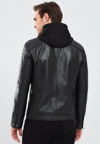 Chaqueta de cuero negra con un diseño ajustado, que cuenta con una capucha negra desmontable, detalles acanalados en los hombros y broches en los puños.