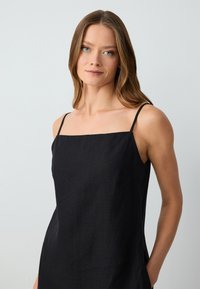 Robe en lin noir avec un décolleté carré et des bretelles étroites. Elle présente une coupe décontractée et des poches latérales, mettant en valeur une texture subtile.