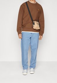 Brauner Sweatshirt, hellblaue lockere Jeans und weiße Sneakers. Kleine beige Umhängetasche mit schwarzen Akzenten. Lässiger und entspannter Schnitt.