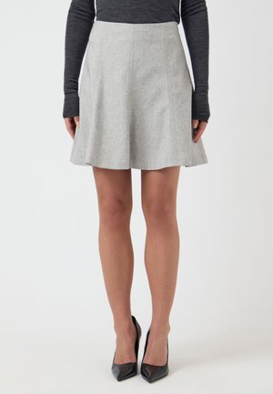 Kobieta w jasnoszarej spódnicy w kształcie litery A, ciemnej bluzce z długim rękawem i czarnych czółenkach na wysokim obcasie z ostrym noskiem stojąca na jednolitym tle.