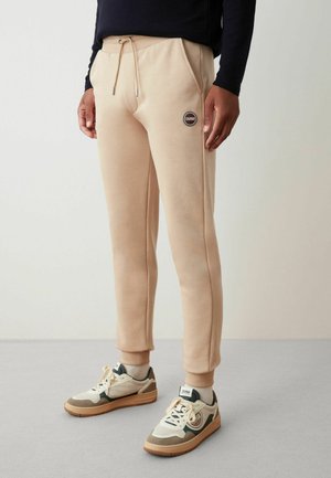 Pantaloni jogger beige con vita elastica, cordino, tasche laterali e patch con logo. Indossati con sneakers color crema e marrone.