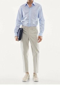 Homme portant une chemise bleu clair à boutons, un pantalon habillé beige et des baskets beiges, tenant une pochette noire dans sa main gauche.