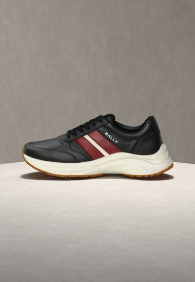 Baskets en cuir noir avec des bandes latérales rouges et blanches, semelle épaisse blanche, semelle extérieure marron, et logo "BALLY" sur le panneau latéral.