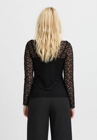 Schwarzes Spitzenoberteil mit langen Ärmeln, floralem Muster, taillierter Silhouette und gewelltem Saum, getragen über schwarzen Hosen. Strukturiert, semitransparenter Stoff.