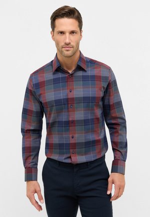 Man met een getailleerd overhemd met lange mouwen in blauw, rood en grijs ruitpatroon, met donkere broek tegen een eenvoudige lichte achtergrond.