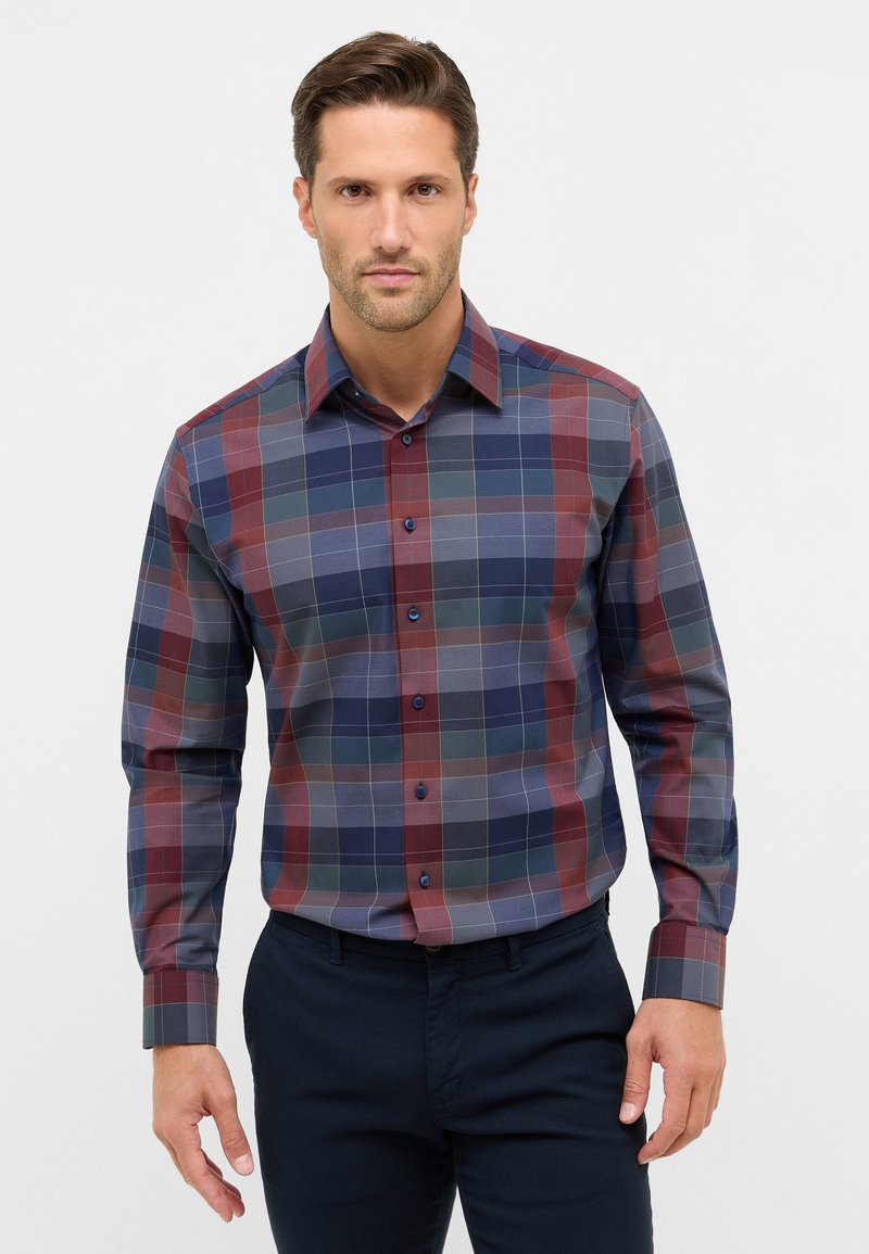 Uomo che indossa una camicia a maniche lunghe aderente con bottoni in una fantasia a quadri blu, rossa e grigia, abbinate a pantaloni scuri, su uno sfondo chiaro e semplice.