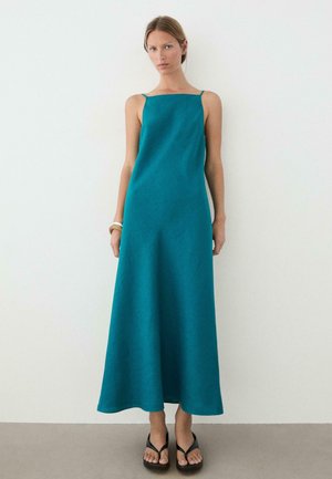 Frau trägt ein ärmelloses Maxikleid in Petrol mit dünnen Trägern, schwarze Plateau-Sandalen und goldene Armreifen, steht vor einer weißen Wand.