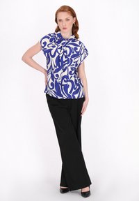 Kurzärmlige Bluse in Creme mit blauem abstrakten Muster, kombiniert mit schwarzen weiten Hosen und schwarzen High Heels. Glatte Stoffqualität, lässige Passform.