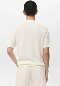 Krátký rukáv, svetr s kulatým výstřihem v krémové barvě, s žebrovanou texturou a přiléhavým střihem, navržený v čistém, minimalistickém stylu.