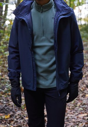 Personne portant une veste bleue, un pull vert à demi-fermeture éclair, des gants noirs et un pantalon noir, debout dans une forêt avec des feuilles d’automne au sol.