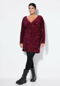 Burgundské šaty se zaměřením na aplikaci flitrů, s obvazovým designem a dlouhými rukávy, spárované s černými neprůhlednými punčocháči a masivními černými botami.