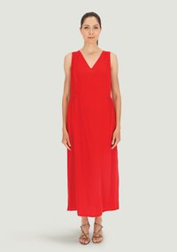 Czerwona, bezrękawnikowa sukienka maxi z dekoltem w kształcie litery V, zwiewna sylwetka oraz fałdy w talii; model nosi sandały na paseczkach. Gładki, lekki materiał.