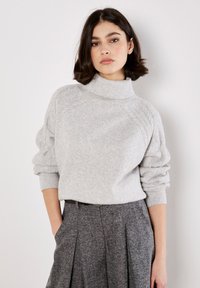 Maglione dolcevita grigio chiaro con dettagli testurizzati sulle spalle e polsini larghi, abbinato a pantaloni grigi scuri plissettati.