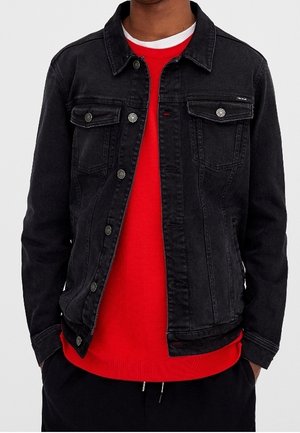 Personne portant une veste en jean noire par-dessus un pull rouge vif et un pantalon noir, debout les mains dans les poches sur un fond uni.