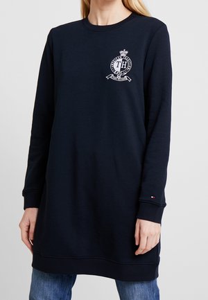 Vrouw draagt een marineblauwe lange mouwen sweatshirtjurk met een klein wit schildlogo op de borst en een klein vlagembleem op de mouwboord.