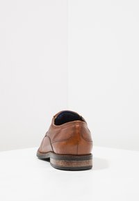 Chaussure en cuir marron avec une texture lisse, dotée de détails perforés, d'un bout arrondi et d'une semelle marron foncé contrastante.