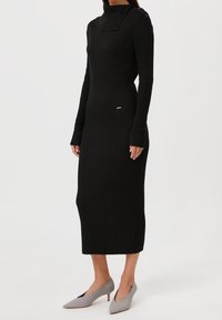 Robe midi noire à côtes avec un col haut, des manches longues et des boutons sur le côté. Portée avec des escarpins pointus gris clair.