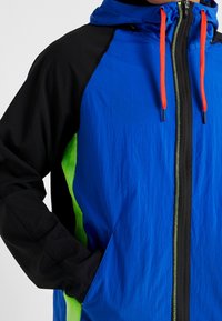 Blå och svart jacka med gröna sidopaneler, framdragkedja och orange snoddar, en hand i fickan, visad från bröstet till midjan.