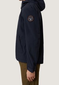 Man i marinblå jacka med huva och märke på överarmen, står sidledes med avslappnad hand vid sidan, mot en enfärgad bakgrund.