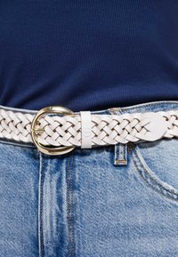 Valkoinen punottu nahkavyö, jossa on kultainen pyöreä solki, käytetty sinisten farkkujen kanssa; vyössä näkyvät ompeleet ja sileä pinta.