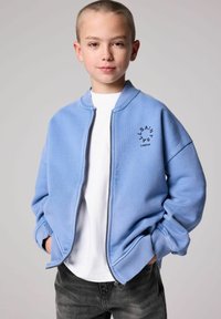 Chaqueta bomber azul claro con cremallera, puños y cuello de canalé. Presenta un logo impreso en el pecho; se lleva sobre una camiseta blanca y jeans.