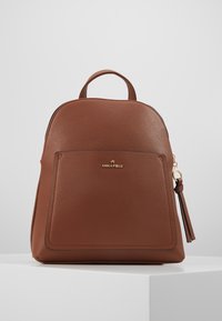 Zaino in pelle marrone con parte superiore arrotondata, tasca frontale, dettagli in metallo oro e un accessorio a nappina. Superficie strutturata con cuciture visibili.