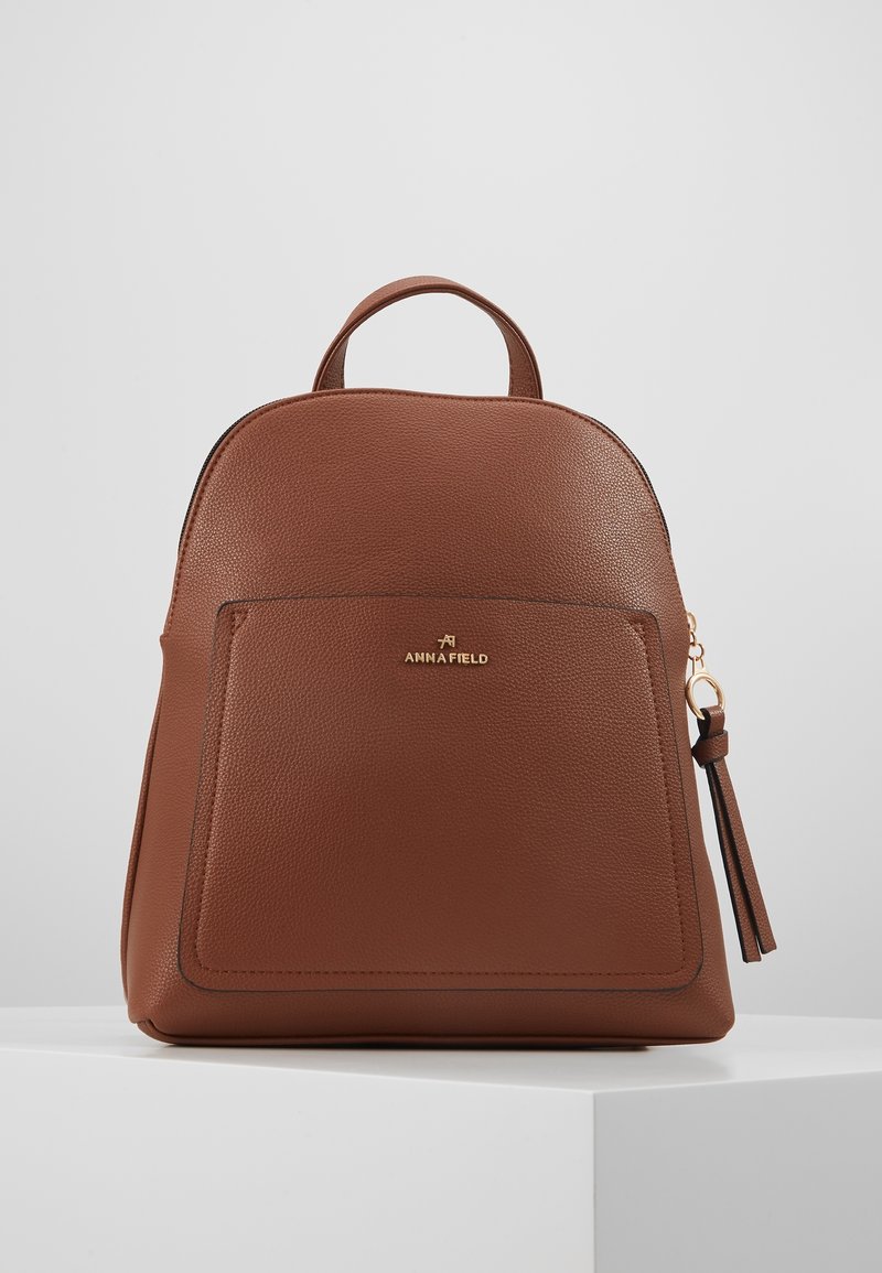 Zaino in pelle marrone con parte superiore arrotondata, tasca frontale, dettagli in metallo oro e un accessorio a nappina. Superficie strutturata con cuciture visibili.