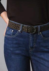 Pasek w leopardzi wzór w ciemnych odcieniach z okrągłą metalową klamr�ą, noszony z ciemnoniebieskimi dżinsami, które mają kieszenie z przodu i detale w stichingu.