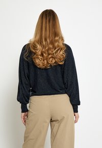 Maglione nero scintillante con maniche lunghe drappeggiate e orlo a coste, abbinato a pantaloni beige chiaro a gamba larga. Tessuto liscio, vestibilità casual.