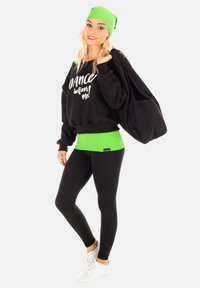 Schwarzer Pullover mit weißem Text, grünem Bund, schwarze Leggings und weißen Sneakern. Grüner Beanie und große schwarze Tasche. Sauberer, sportlicher Look.