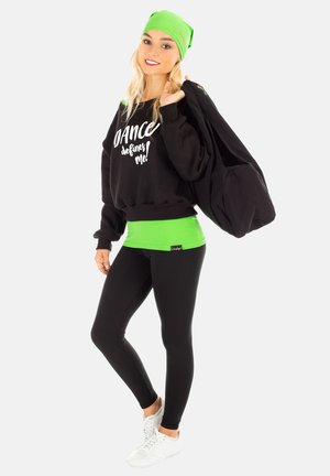 Schwarzer Pullover mit weißem Text, grünem Bund, schwarze Leggings und weißen Sneakern. Grüner Beanie und große schwarze Tasche. Sauberer, sportlicher Look.