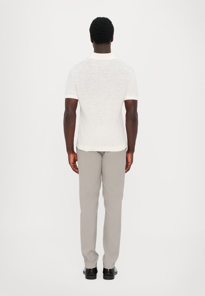 Polo-shirt blanc à manches courtes avec une finition texturée, associé à un pantalon gris clair et des chaussures noires, vu de derrière.