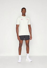 Svijetlosiva sportska majica kratkih rukava s ružičastim logotipom. Kombinirana s crnim kratkim hlačama i bijelim tenisicama, stoji ispred neutralne pozadine.