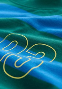 Nahaufnahme eines grün-blau gestreiften Stoffes mit gelb umrandeten Zahlen "02", die darauf gedruckt sind.
