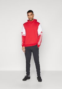 Röd sweatshirt med vita ärmdetaljer och tre vita ränder på axlarna, matchad med mörkgrå joggers och svarta skor.