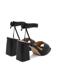 Sandali neri con tacco spesso e cinturini annodati alla caviglia, realizzati in pelle liscia con un design open toe.