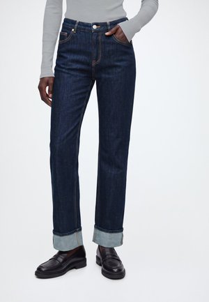 Personne portant un jean bleu foncé retroussé, un haut gris à manches longues et des mocassins noirs, debout avec une main dans la poche.
