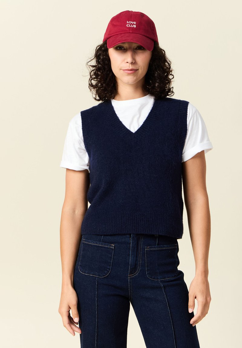 Mörkblå stickad väst med V-formad halsringning ovanpå en vit T-shirt, kombinerad med mörka högmidjade jeans. En röd keps med texten "LOVE CLUB."